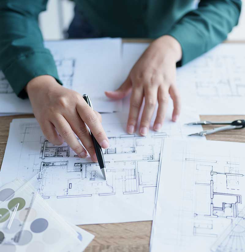 Person in a green shirt reviews and marks up architectural floor plans on a desk, surrounded by drafting tools, design samples, and premium tile Florida catalogs for luxury surface specification.
