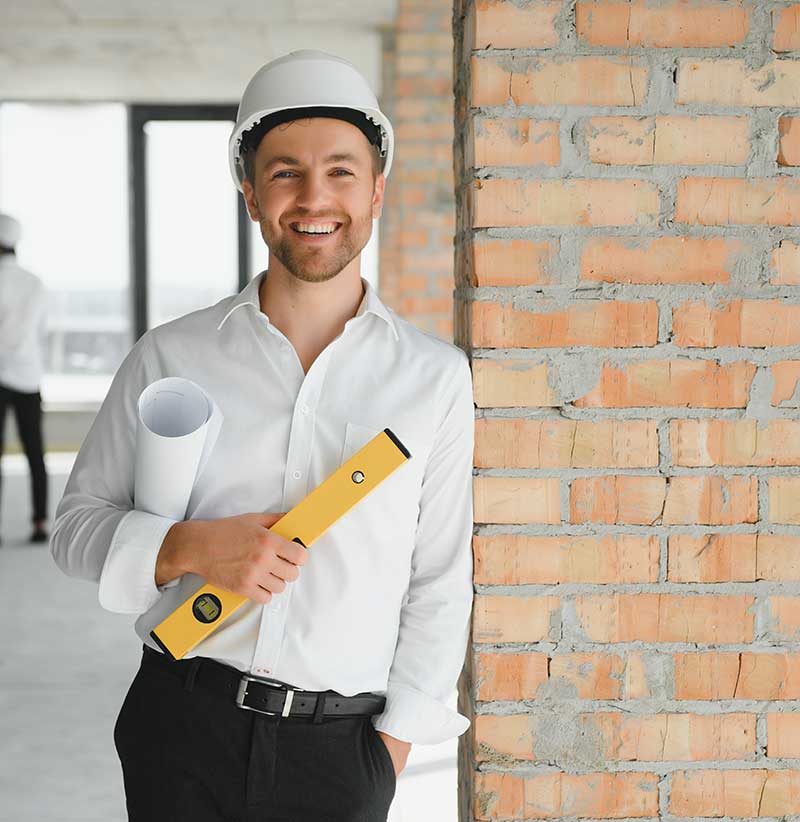Man wearing a white hard hat and holding blueprints and a yellow level, standing by an unfinished brick wall at a construction site, consulting on luxury surface specification as an expert tile and stone consultant.