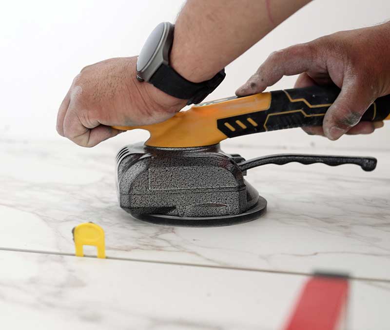 A person, possibly a tile and stone consultant, uses a handheld tool with a yellow handle and circular base on a white marble surface, with a yellow spacer nearby.